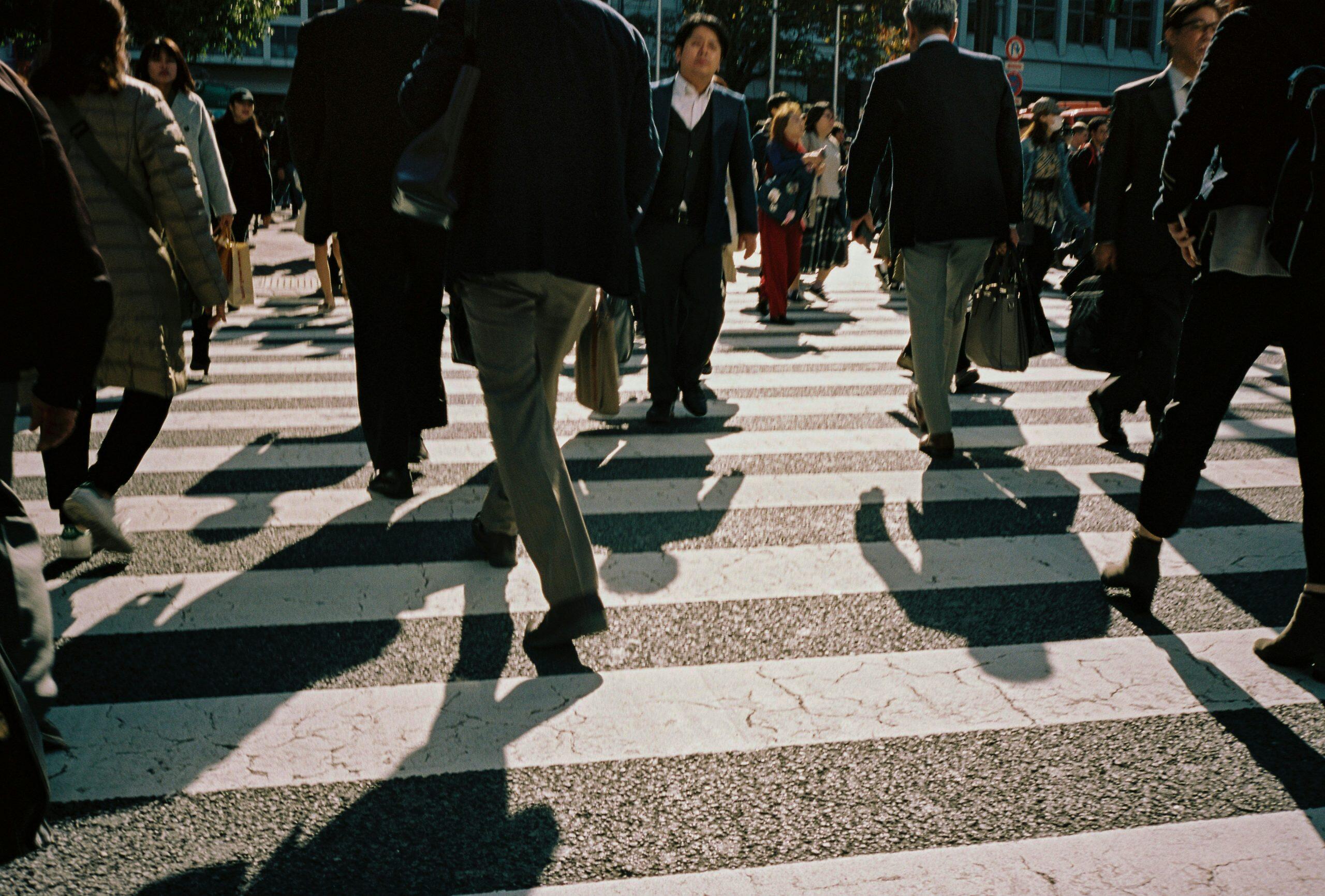 A busy pedestrian crossing in a city with people walking in various directions, dressed in a mix of business and casual attire, casting long shadows on the zebra crossing under angled sunlight, with trees and buildings in the background, reminiscent of the energy seen during conferences in Las Vegas this week.