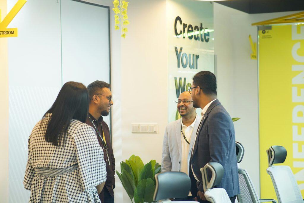 Four people engaged in a semi-circle conversation in a modern office with yellow and white decor, indoor plants, ergonomic chairs, and visible signage reading 'ROOM' and 'Create Your World' on the walls, including a marketing strategist contributing ideas.