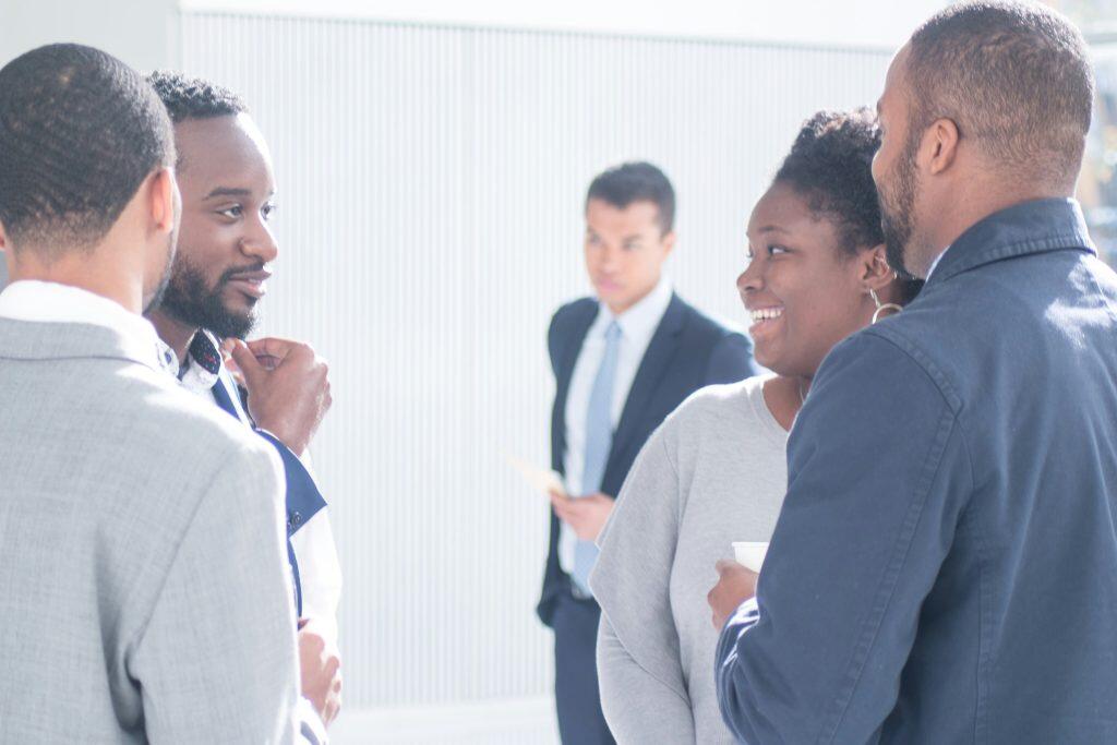 Five individuals at a professional event, with four in the foreground engaged in conversation, dressed in business attire, and one person in the background holding a paper and observing, set in a bright, well-lit environment at conferences in Las Vegas this week.