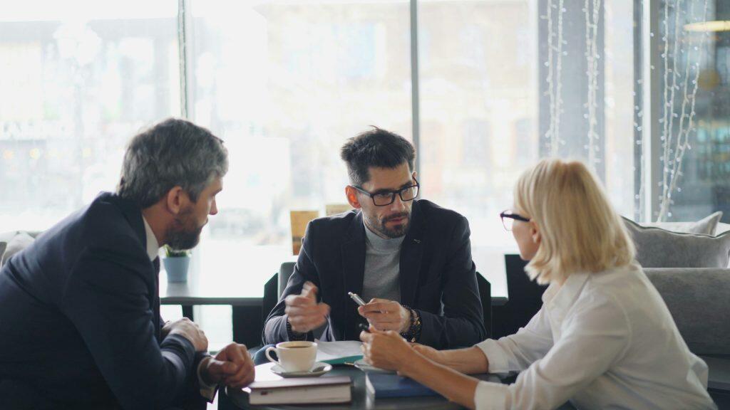 Three people seated around a small table in a modern setting, engaged in a collaborative discussion, with one person gesturing with a pen, a cup of coffee, and documents on the table, and a plant visible in the background. One person appears to be a marketing strategist, contributing to the conversation.